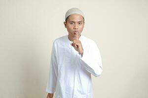 porträtt av attraktiv asiatisk muslim man i vit skjorta viskande illvillig prata konversation, hand på mun talande hemlighet rykte. isolerat bild på grå bakgrund foto