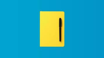 Foto av stängd gul anteckningsbok och kulpenna penna över blå bakgrund. minimalistisk bild av stängd dagbok och svart penna som tillbaka till skola begrepp.
