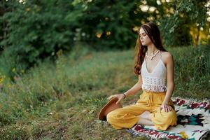 en ung hippie kvinna mediterar i natur i de parkera, Sammanträde i en lotus placera på henne färgrik pläd och njuter harmoni med de värld i eko-kläder foto