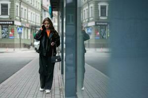 ung lady i läder jacka gående i urban stad, tala på telefon. foto