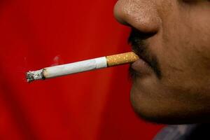 rökare med en cigarett i hans mun på en suddigt röd bakgrund. ohälsosam livsstil med en manlig rökare. man rökning närbild se. cigarett brinnande i en rökare mun. manlig rökare med en skägg. foto