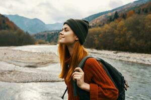 Lycklig resande med en ryggsäck i en hatt och en Tröja i de bergen nära de flod foto