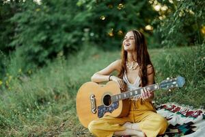 ung kvinna hippie konstnär spelar gitarr och sjunger låtar i miljövänlig Kläder Sammanträde på de jord utanför i natur i de höst ser ut på de solnedgång foto