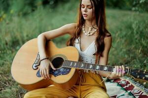 hippie kvinna leende och kramas henne gitarr i natur i de parkera i de solnedgång ljus i eko-kläder foto