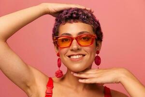 mode porträtt av en kvinna med en kort frisyr i färgad solglasögon med ovanlig Tillbehör med örhängen leende på en rosa ljus bakgrund foto
