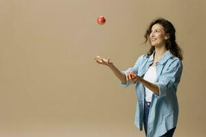 Lycklig leende fokuserade lockigt skön kvinna i jeans tillfällig skjorta jonglering med äpple Framställ isolerat på över beige pastell bakgrund. friska mat. naturlig miljövänlig Produkter begrepp. kopia Plats foto