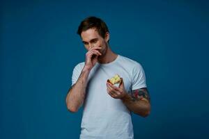 söt kille med en tatuering och i en vit t-shirt på en blå bakgrund äter äpplen beskurna se foto