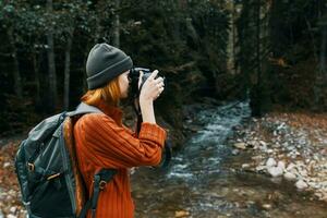 kvinna fotograf innehav en kamera i hand nära de flod i de bergen och skog i de bakgrund foto