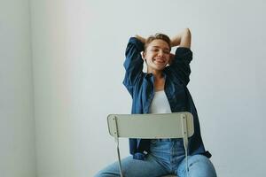 en ung kvinna Sammanträde i en stol på Hem leende med tänder med en kort frisyr i jeans och en denim skjorta på en vit bakgrund. flicka naturlig poser med Nej filter foto