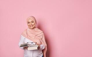 Lycklig muslim gravid kvinna i rosa hijab, leende ser på kamera, innehav glasögon och bok. islam och utbildning foto