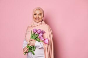 skön muslim gravid bär en rosa hijab, innehav en bukett av lila tulpaner för mors dag, leende på kamera foto
