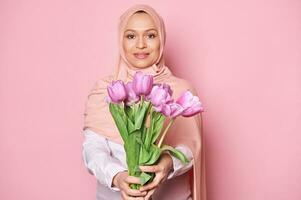 detaljer på en skön bukett av lila tulpaner i de händer av suddig muslim kvinna i rosa hijab, leende på kamera foto