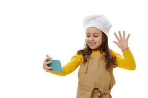 leende positiv liten barn flicka i vit kockens hatt och beige kök förkläde vinka med hand under video konferens foto