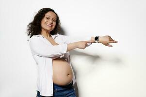 Lycklig kvinna i 28 vecka av graviditet, stående bar mage, som visar visa av henne smart klocka på isolerat vit bakgrund foto