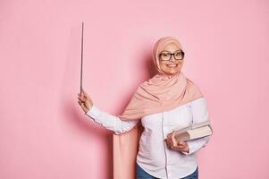 glad erfaren mitten åldrig arab musim lärare i rosa hijab, poäng med pekare på kopia Plats på rosa bakgrund foto