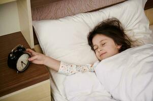 sovande liten flicka i pyjamas, liggande i mysigt säng, växlande av de ringande larm klocka och vakna upp i de morgon- foto