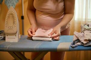 närbild gravid kvinna, de förväntans mor veck strykas kläder för nyfödda, förbereder väska för de moderskap sjukhus foto