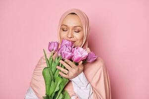 närbild skön muslim kvinna i rosa hijab, innehav en bukett av tulpaner, Framställ med henne ögon stängd, rosa bakgrund foto