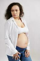 självsäker lugn gravid kvinna, bär knäppte upp vit skjorta och blå denim, ser åt sidan, isolerat vit bakgrund foto