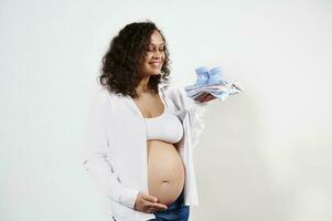 mång etnisk härlig gravid kvinna innehav kläder för henne nyfödd bebis, försiktigt strök henne mage, vit bakgrund foto
