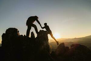 silhuett av lagarbete av två män vandrare portion varje Övrig på topp av berg klättrande team. lagarbete vänskap vandring hjälp varje Övrig förtroende bistånd silhuett i berg, soluppgång. foto