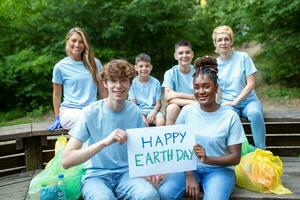 Lycklig volontärer innehav plakat med 'Lycklig jord dag' meddelande. volontärarbete, välgörenhet, rengöring, människor och ekologi begrepp - grupp av Lycklig volontärer med sopor påsar rengöring område i parkera foto