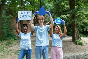 Lycklig volontärer innehav plakat med 'Lycklig jord dag' meddelande. volontärarbete, välgörenhet, rengöring, människor och ekologi begrepp - grupp av Lycklig volontärer med sopor påsar rengöring område i parkera foto