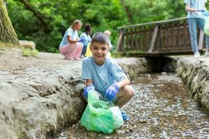 liten pojke plockar upp några återvinning från de flod. familj samlar återvinning och sätta den i en grön väska. mor undervisning barn handla om hållbarhet. foto
