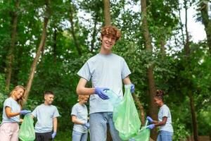 porträtt av ung man med små grupp av volontärer på bakgrund med handskar och sopor påsar rengöring upp stad parkera - miljö bevarande och ekologi begrepp. Allt bär en blå t-tröjor foto