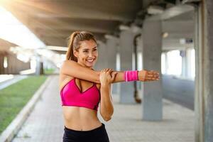 ung sportig kvinna Träning. kvinna håller på med träna övningar på gata. skön atletisk passa flicka i ljus sporter Kläder stretching henne ärm och avkopplande efter kondition Träning på gata. foto