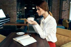 skön kvinna med en kopp i hand sitter på en tabell i en Kafé och bok en restaurang foto