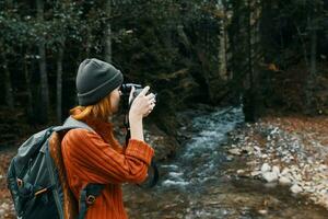 kvinna fotograf innehav en kamera i hand nära de flod i de bergen och skog i de bakgrund foto