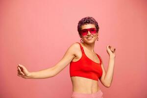 mode porträtt av en kvinna med en kort frisyr i färgad solglasögon med ovanlig Tillbehör med örhängen leende på en rosa ljus bakgrund med en kondition kropp dans foto