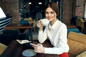 skön kvinna med en kopp i henne hand sitter på en tabell i en Kafé och en bok i henne hand restaurang modell foto