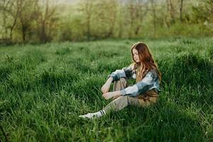 en ung, skön kvinna lounger på de grön gräs i de parkera bär gymnastiksko byxor och en pläd skjorta och utseende ut in i de miljö Sol foto