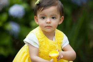 ljuv ett år gammal bebis flicka klädd i gul vattning de växter på de trädgård foto