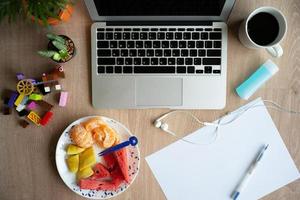 ovanifrån av en bärbar dator på träskrivbordet med stillastående, kaffe och blandad frukt på en vit tallrik. arbeta hemifrån. karantän hemma foto