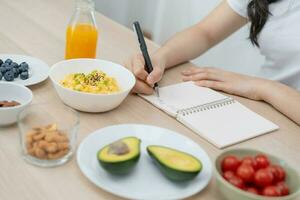 diet, bantning hand av asiatisk ung kvinna, flicka skriva diet planen näring på tabell är annorlunda mat Ingredienser i de grön. dietist av friska, näring av vikt förlust, hälsa vård människor. foto