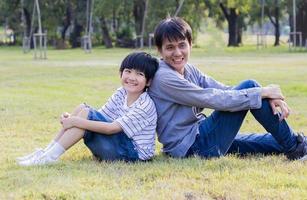 asiatiska far och son sitter glatt på gräsmattan i parken foto