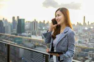 ung asiatisk företag kvinna i formell kostym är telefon kallelse kund medan stående utanför de skyskrapa byggnad för marknadsföring, förbindelse företags- arbete, verklig egendom, hus och urban utveckling foto