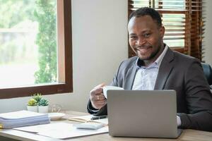 afrikansk amerikan affärsman i en svart kostym innehav en kopp av kaffe till koppla av efter kontroll företag arbete på en bärbar dator inuti de kontor. foto