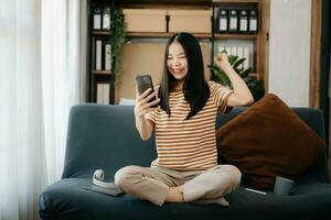 attraktiv Lycklig ung asiatisk studerande studerar på de högskola bibliotek, Sammanträde på de soffa, använder sig av en bärbar dator dator, läsplatta och hörlurar har en video chatt. foto