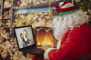 Lycklig santa claus gör video ring upp litet barn talande till barn, hälsning glad jul, Lycklig ny år i virtuell video uppkopplad chatt på bärbar dator, Sammanträde i främre av de öppen spis. foto