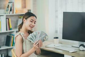 asiatisk kvinna flicka leende och innehav dollar räkningen på trä tabell skrivbord i levande rum på Hem. sparande investering rikedom begrepp. foto