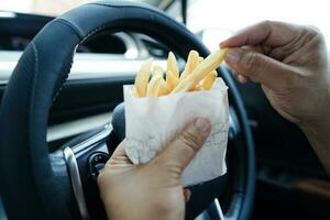 asiatisk kvinna förare håll och äta franska frites i bil, farlig och risk ett olycka. foto