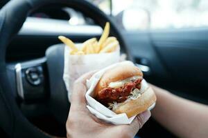 asiatisk kvinna förare håll och äta hamburgare och franska frites i bil, farlig och risk ett olycka. foto