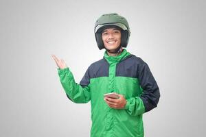 porträtt av asiatisk uppkopplad taxi förare bär grön jacka och hjälm innehav mobil telefon och pekande till tömma Plats med finger. isolerat bild på vit bakgrund foto