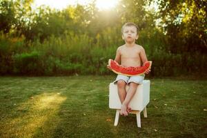Lycklig leende barn pojke äter de vattenmelon utomhus- i de bakgård foto
