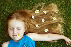 porträtt av en skön liten flicka med friska röd hår med kamomill blommor liggande på de gräs foto