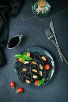 klar att äta svart spaghetti med musslor, tomater och basilika på en tallrik på en svart bakgrund. mat fotografi i mörk färger. topp och vertikal se foto